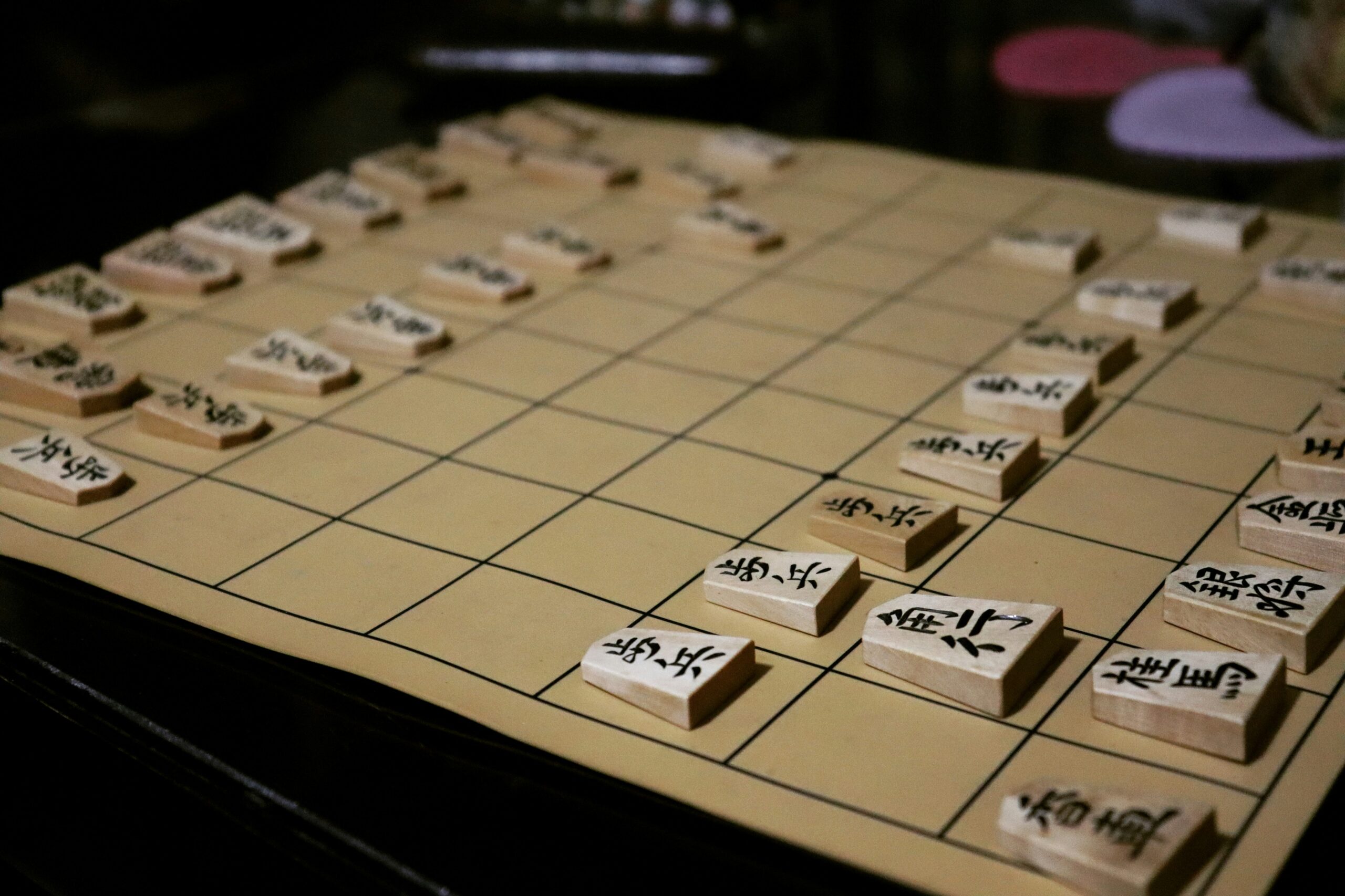 薄暗い照明の中、対局途中の将棋盤に並ぶ木の駒。一手ずつ決断を重ねる将棋は、リーダーの意思決定と「決断疲れ」を象徴するイメージ。