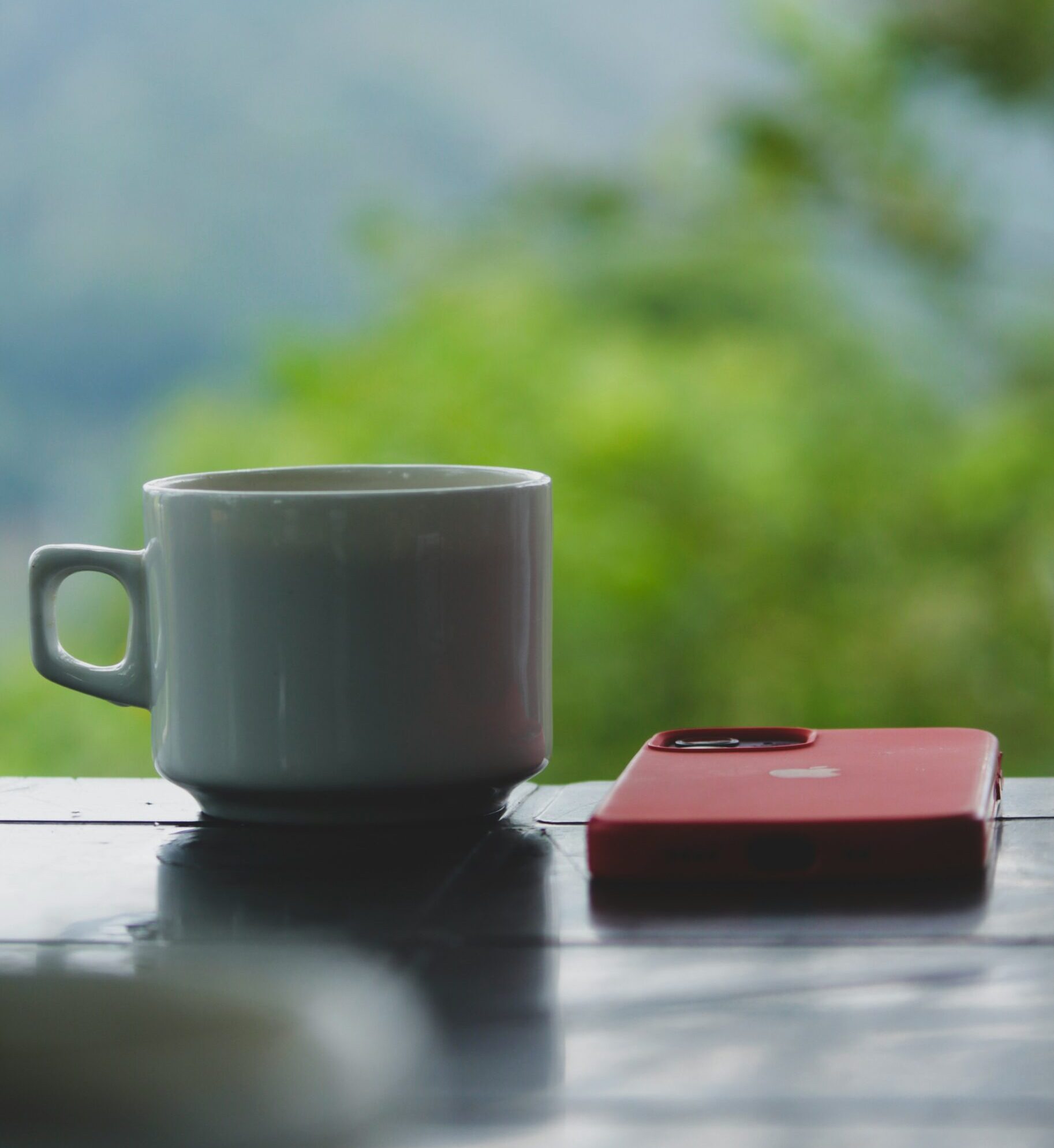 緑の山を背景に、白いコーヒーカップと置かれた赤いスマホ｜SNSから離れて心を休める情景